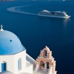 One of the blue domed churches of Santorini on the Caldera with a pink bell tower next to it. In the background is a cruise ship