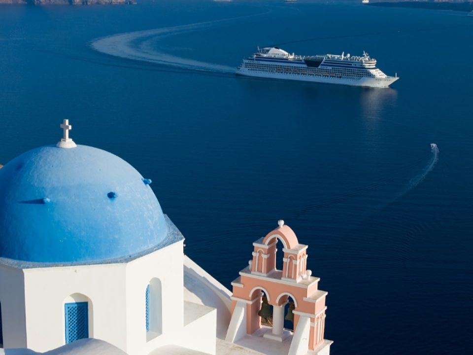 One of the blue domed churches of Santorini on the Caldera with a pink bell tower next to it. In the background is a cruise ship